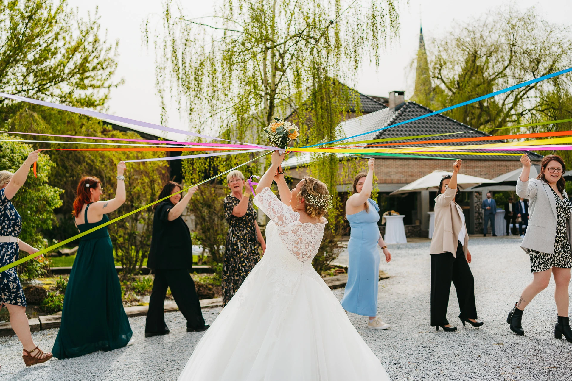 Une mariée en robe blanche et casque fleuri tient des rubans colorés reliés aux invités marchant en cercle lors d'une cérémonie de mariage coordonnée par un organisateur.