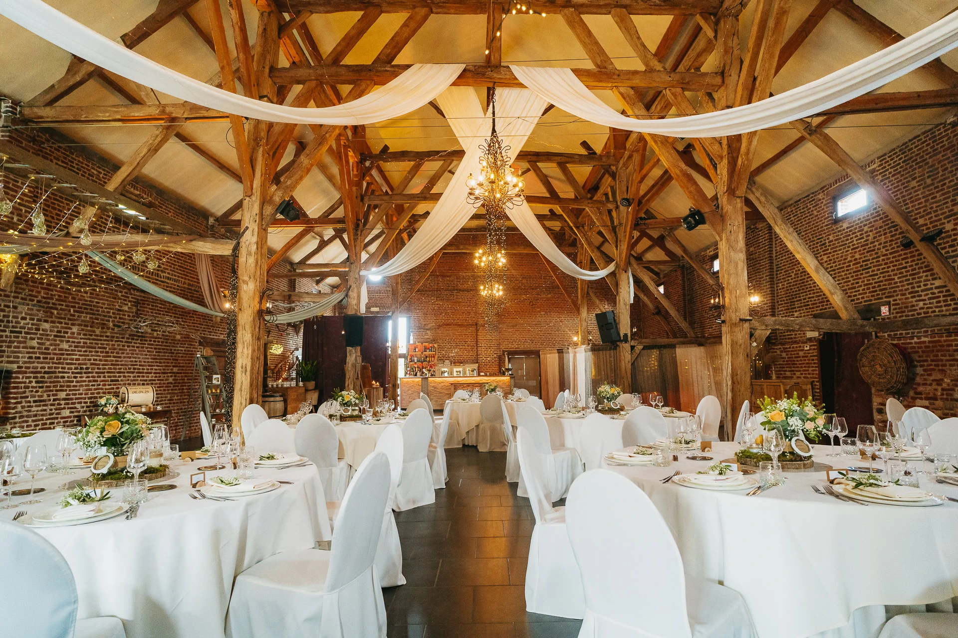 Un lieu de réception de mariage rustique avec poutres apparentes, rideaux blancs, tables rondes et centres de table fleuris, parfaitement coordonné par un organisateur de mariage.
