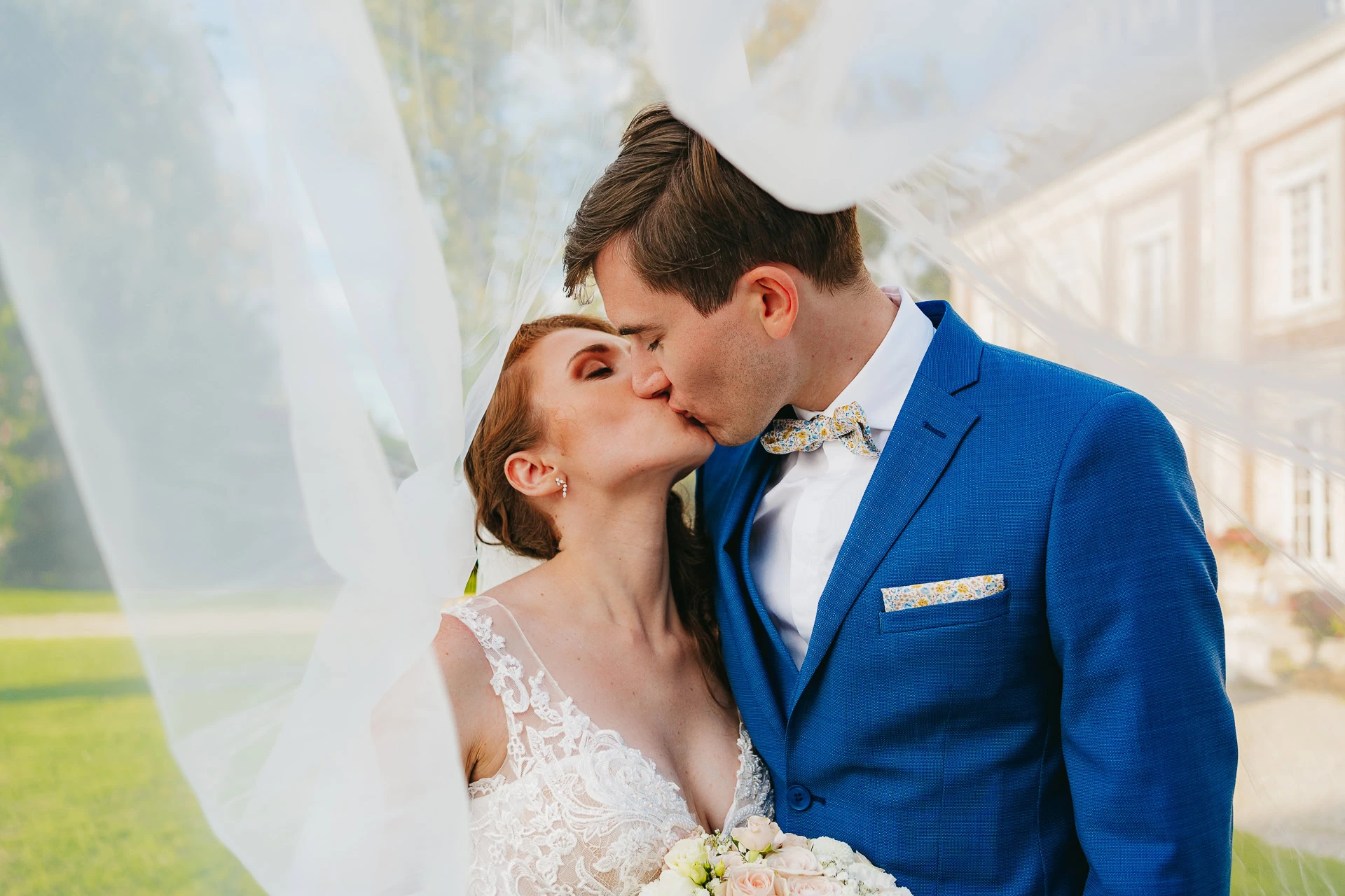 Un baiser des mariés sous un voile gonflé dans un décor extérieur organisé par leur wedding planner. Le marié porte un costume bleu et la mariée une robe blanche avec un bouquet.