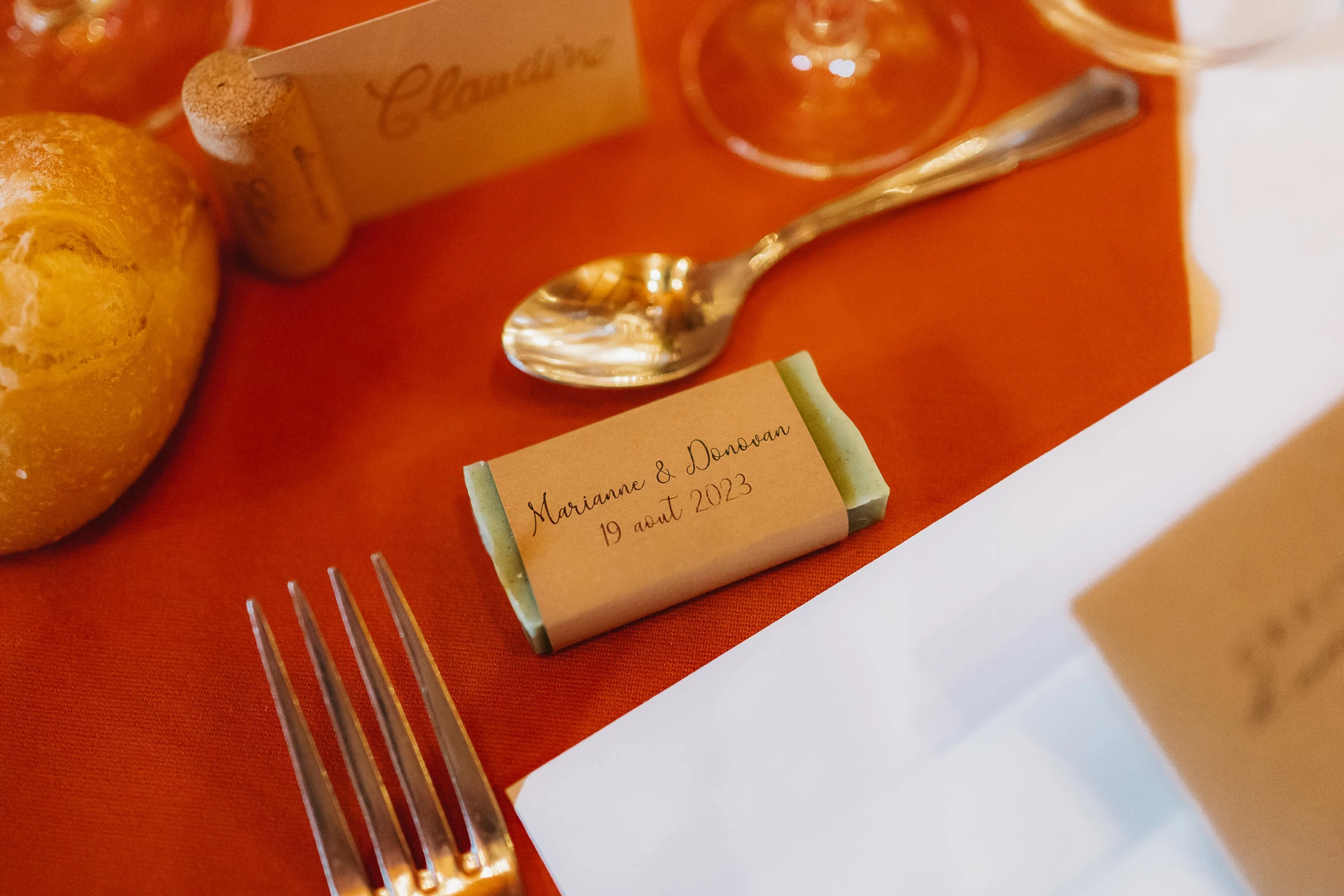 Un couvert avec une étiquette nominative et un petit savon personnalisé emballé avec la date du mariage sur une nappe rouge.
