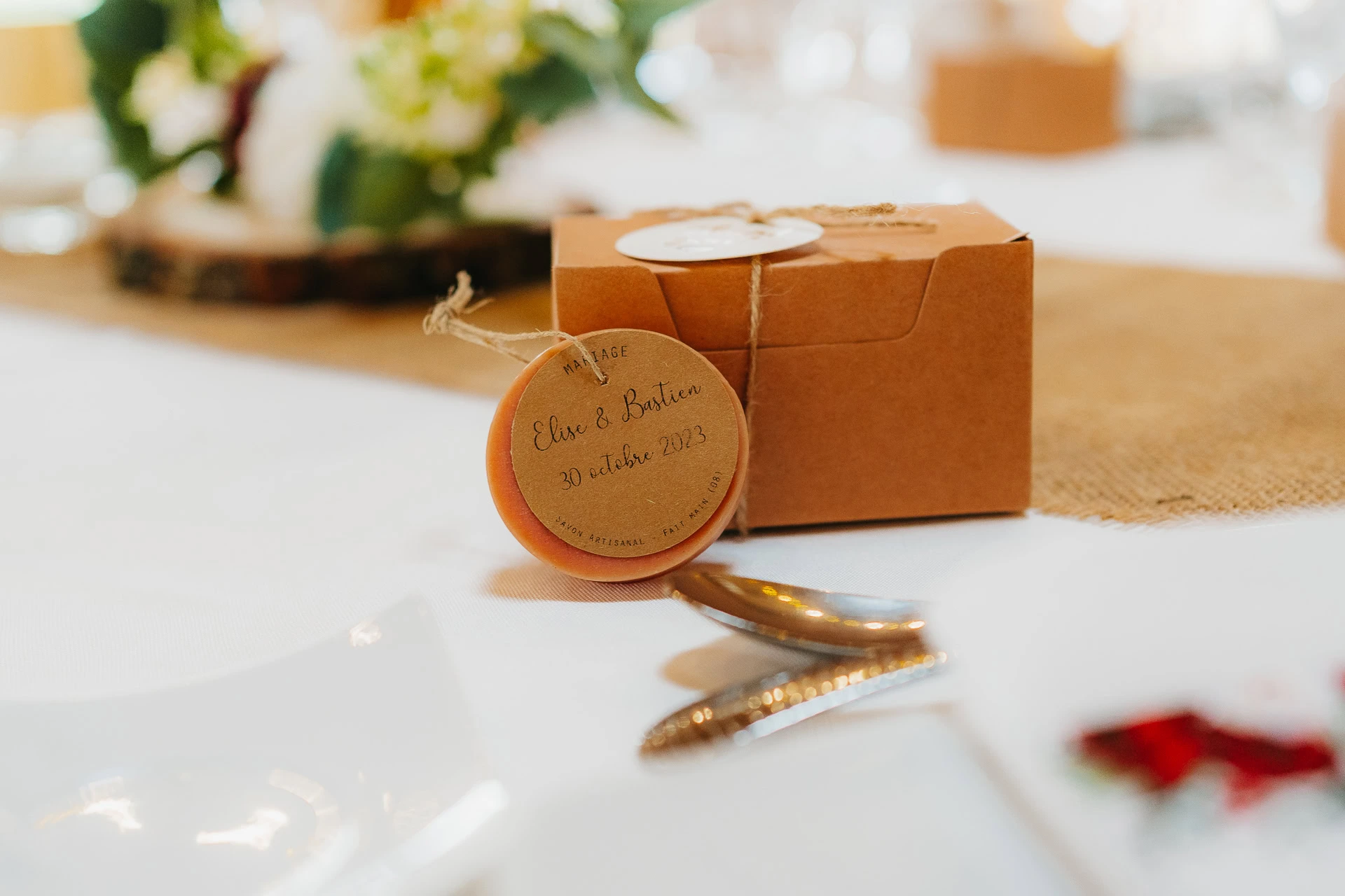 Une petite boîte en carton marron posée sur une table, nouée avec de la ficelle et une étiquette ronde sur laquelle est écrit la date du mariage. Un arrangement floral et une mise de table en arrière-plan.