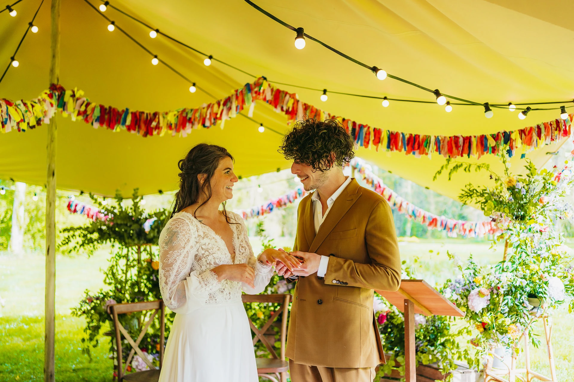 Des mariés se tiennent sous une tente décorée avec des guirlandes lumineuses, se tenant la main et se souriant pendant leur cérémonie de mariage.