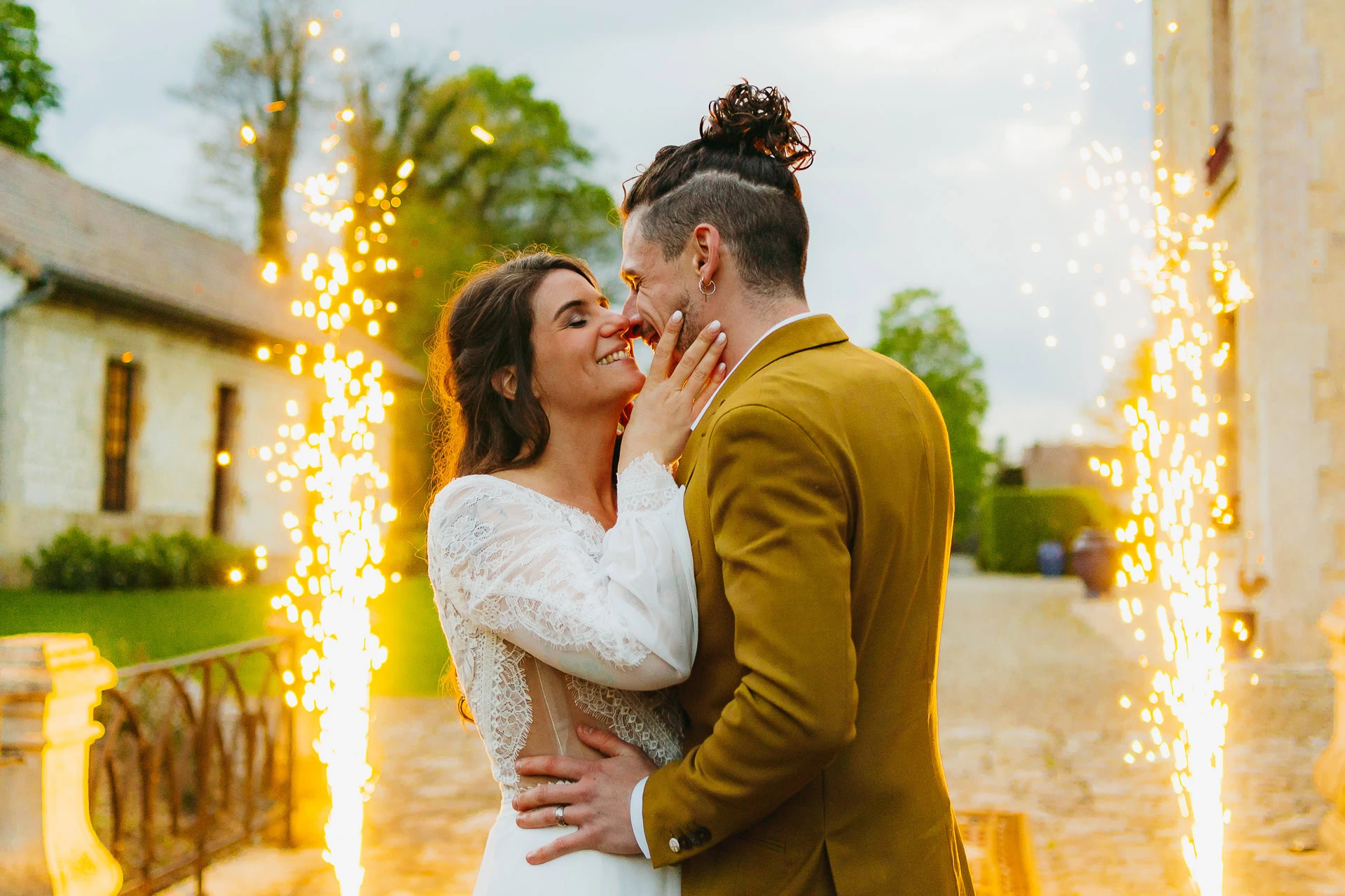 Un couple en tenue de mariage s'embrasse et sourit joyeusement avec des cierges magiques en arrière-plan.