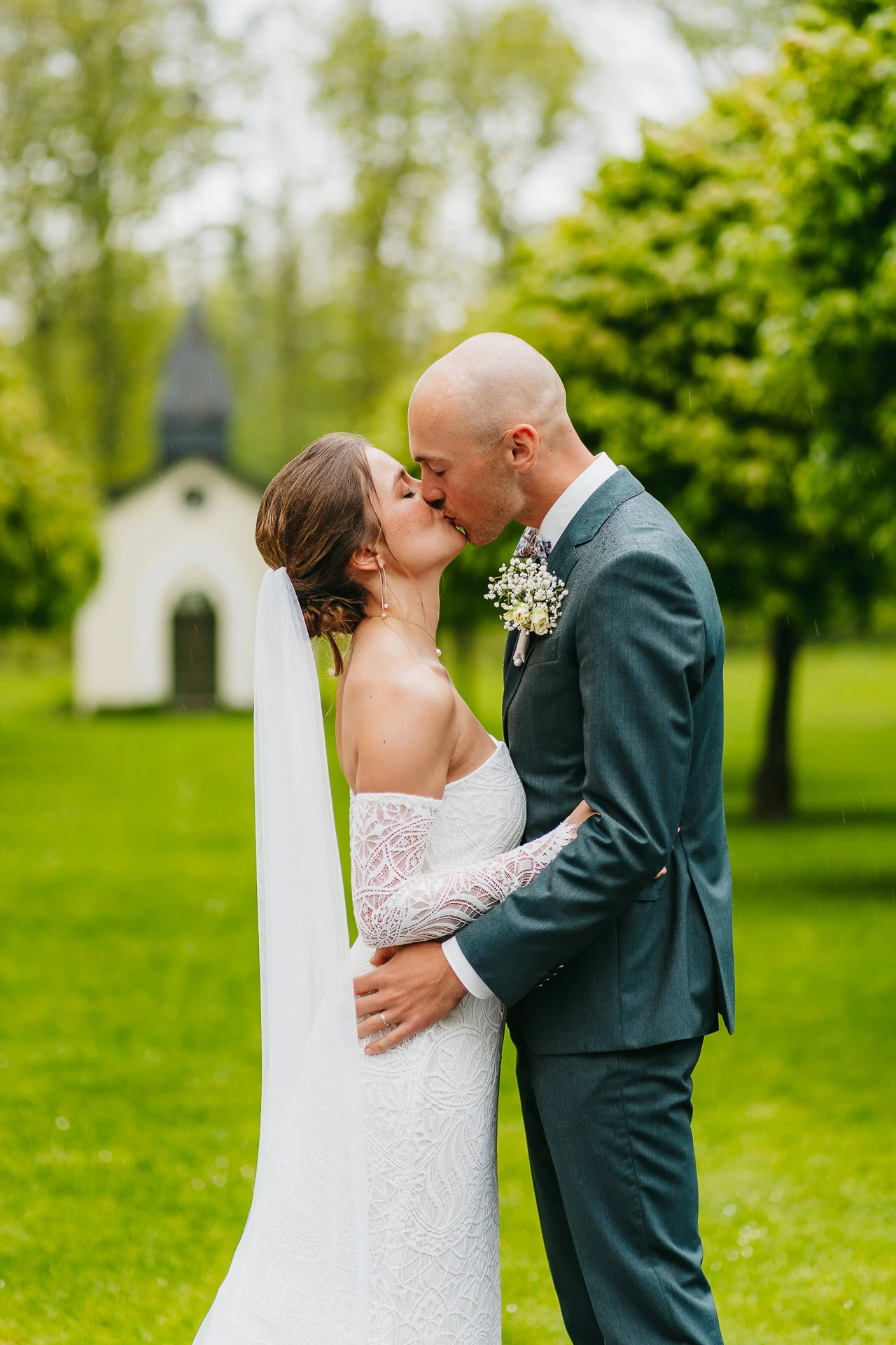 Les mariés partagent un baiser à l'extérieur. La mariée porte une robe blanche et un voile, tandis que le marié porte un costume avec une boutonnière. Ils sont entourés de verdure, avec une petite chapelle en arrière-plan.
