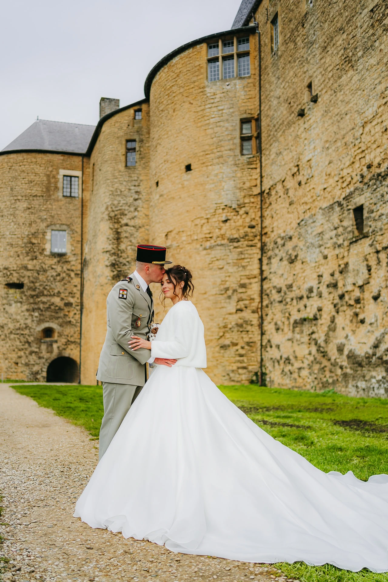 Les mariés partagent un baiser devant un grand château de pierre. Le marié porte un uniforme militaire et la mariée porte une robe de mariée blanche avec une longue traîne.
