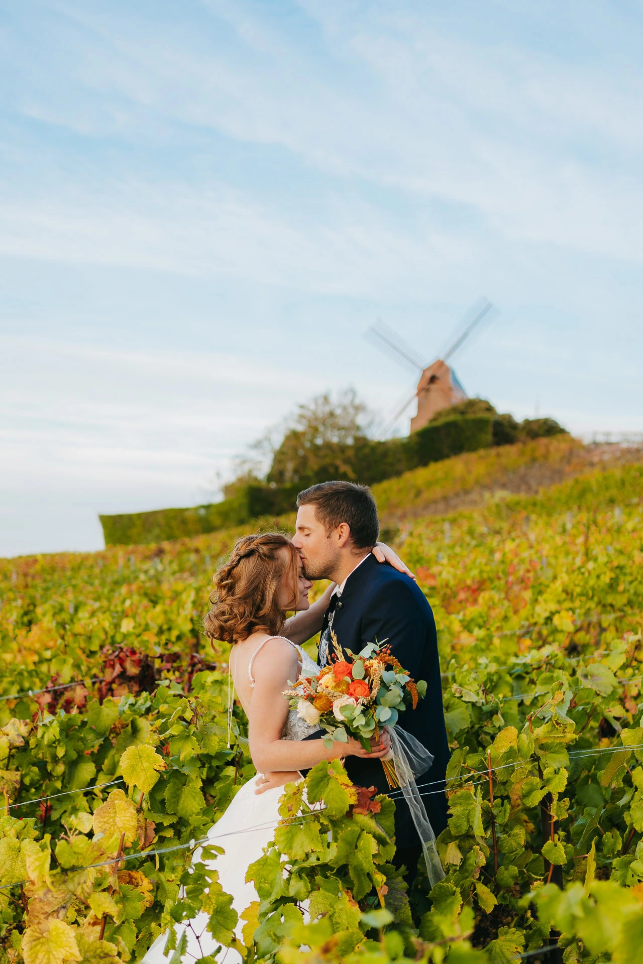 Un couple en tenue de mariage partage un baiser dans un vignoble, avec un moulin à vent en arrière-plan. La mariée tient un bouquet de fleurs.