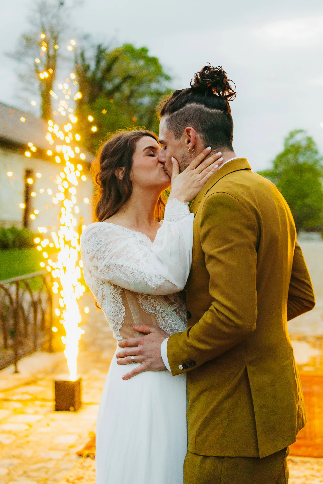 Une mariée en robe blanche et un marié en costume moutarde s'embrassent à l'extérieur devant un petit feu d'artifice.