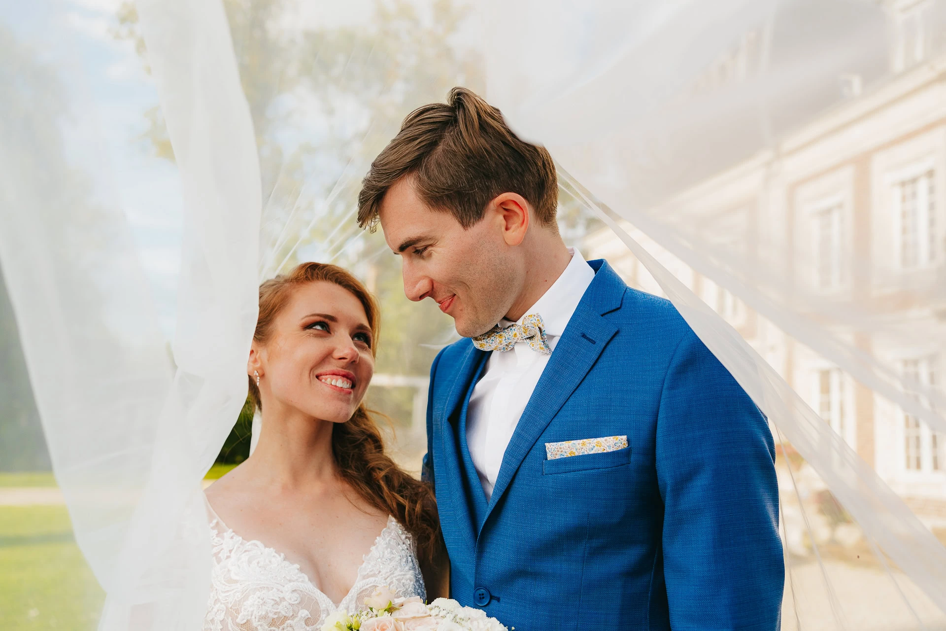 Les mariés se tiennent sous un voile et se regardent. Le marié porte un costume bleu et la mariée une robe en dentelle blanche.